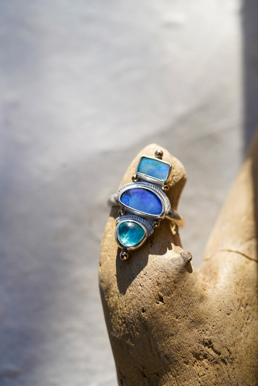 A silver ring with three stacked bright blue stones in vibrant blue and turquoise tones, displayed on a piece of driftwood against a neutral light background.