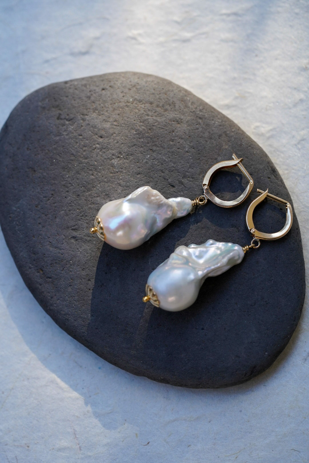 A pair of dangle earrings with irregular white baroque pearls and gold hoops, displayed on a dark gray stone against a light gray textured background.