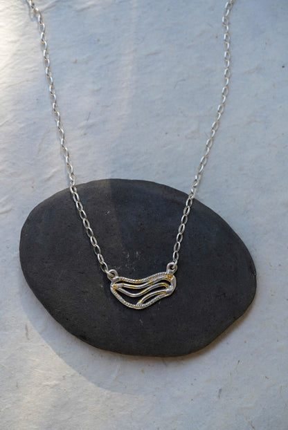 A silver chain necklace with organic shaped pendant featuring textured gold dusting, displayed on a smooth dark gray stone against a light gray background.
