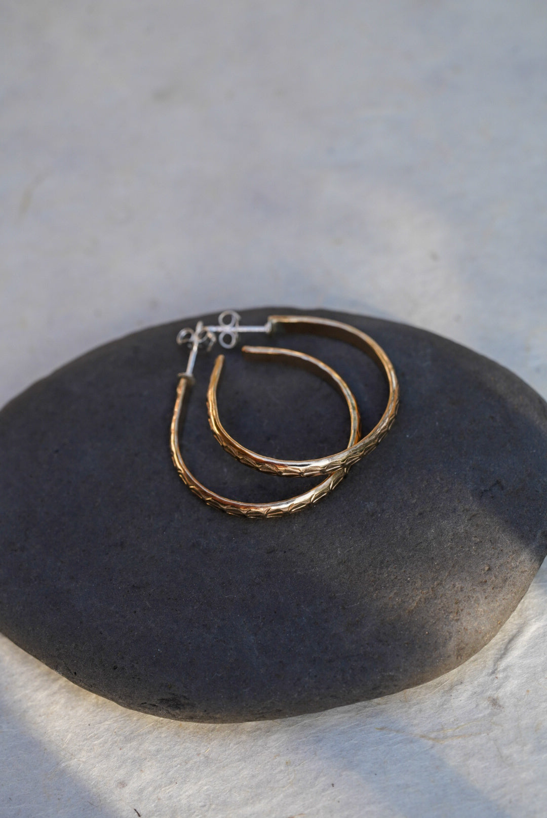 A pair of textured gold hoop earrings resting on a dark gray stone, set against a light gray stone background.