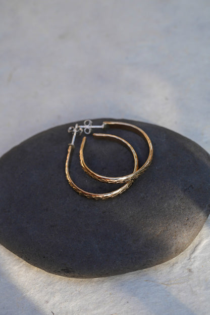 A pair of textured gold hoop earrings resting on a dark gray stone, set against a light gray stone background.