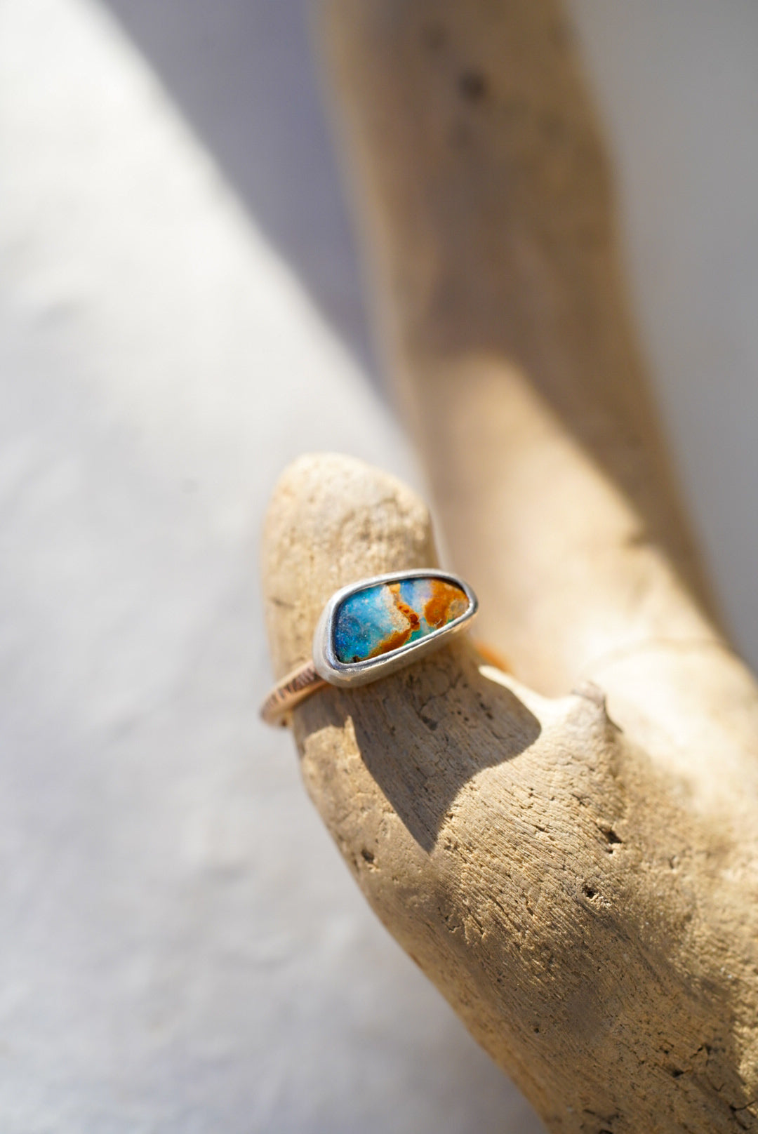 A silver ring with a turquoise and orange opal, displayed on a piece of driftwood against a bright, sunlit white background.
