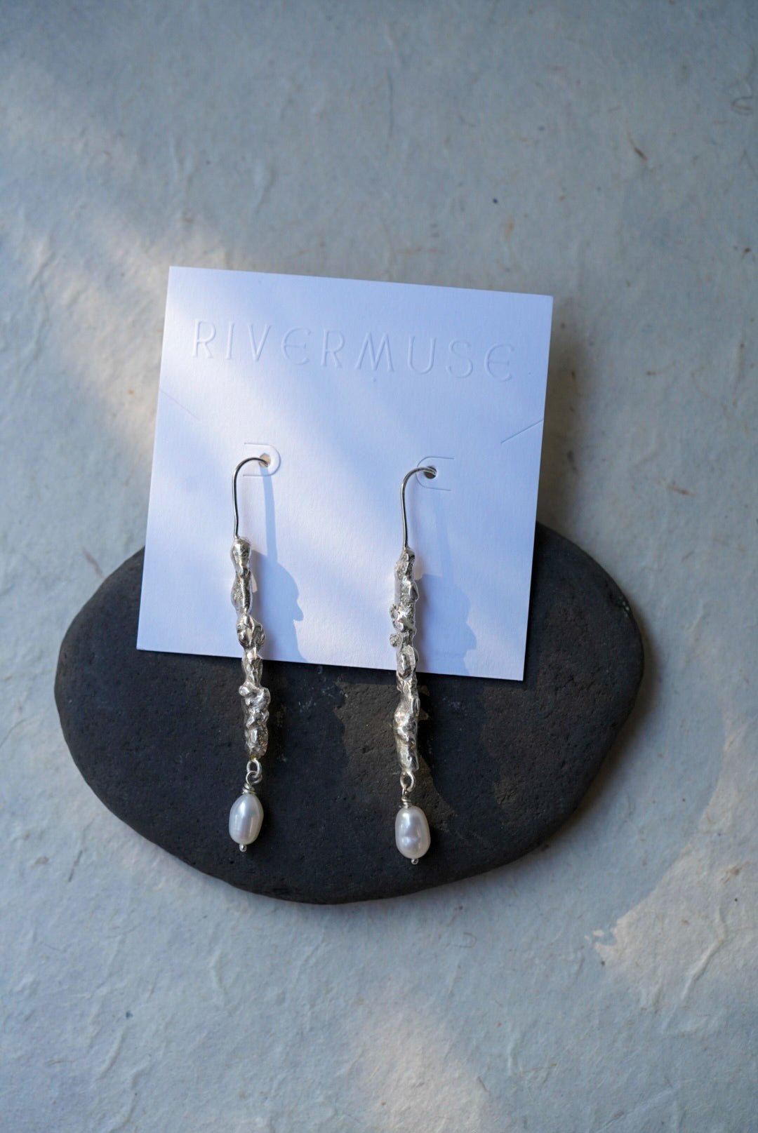 A pair of handmade textured recycled silver dangle earrings and small white pearl-like drops, displayed on a white card labeled RIVERMUSE resting on a dark stone against a light textured wall