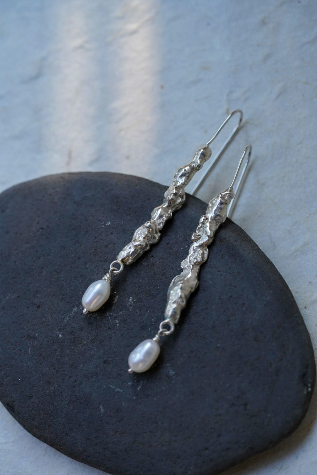 A pair of handmade textured recycled silver dangle earrings and small white pearl-like drops, displayed on a dark oval stone against a light textured wall.