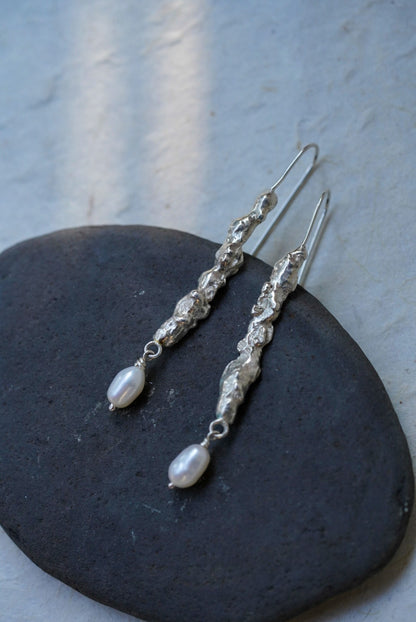 A pair of handmade textured recycled silver dangle earrings and small white pearl-like drops, displayed on a dark oval stone against a light textured wall.
