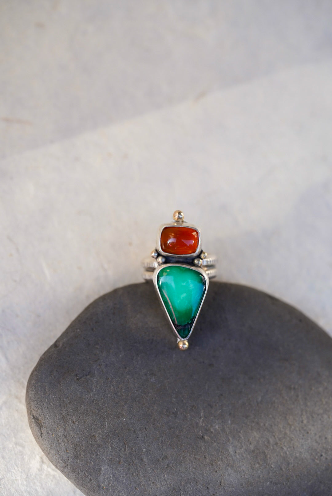 A silver ring featuring a large green teardrop-shaped variscite stone and a smaller orange oval chalcedony, displayed on a dark gray stone against a light gray background.