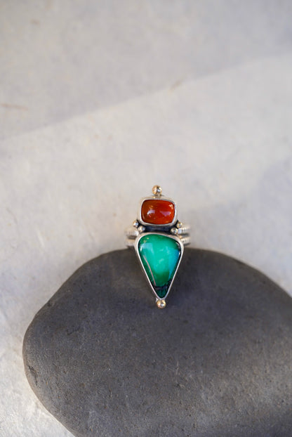 A silver ring featuring a large green teardrop-shaped variscite stone and a smaller orange oval chalcedony, displayed on a dark gray stone against a light gray background.