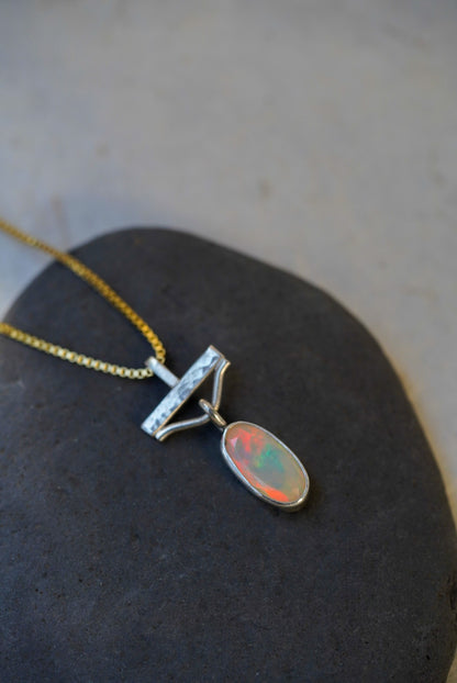 A silver pendant necklace with an oval opal stone showing orange, green, and blue flashes, resting on a dark stone surface.