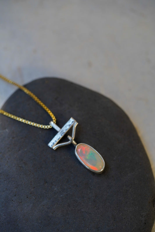 A silver pendant necklace with an oval opal stone showing orange, green, and blue flashes, resting on a dark stone surface.