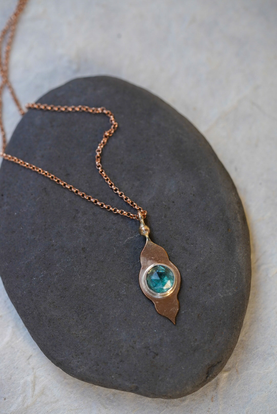 A rose gold necklace with a temple-shaped pendant featuring a round blue-green stone, displayed on a dark slate stone against a light textured background.