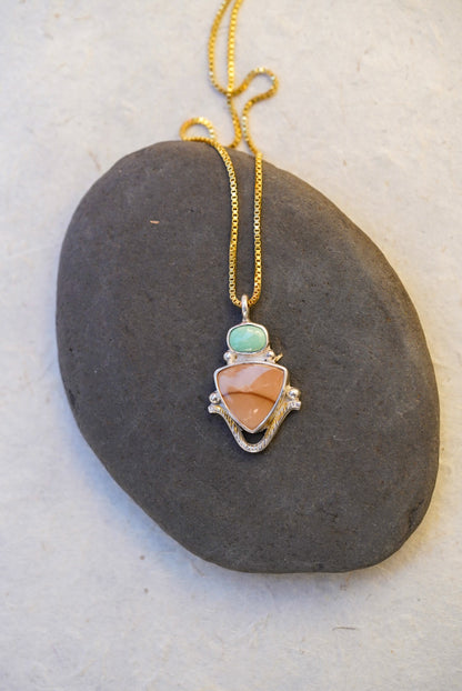 A gold chain necklace with a pendant featuring a light green stone, a neutral jasper stone, and silver metal details, displayed on a dark gray stone against a light textured background.