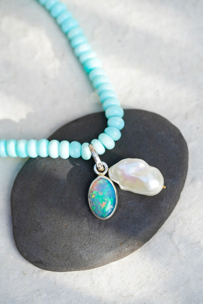 A baby blue beaded necklace with removable charms in rainbow opal and large baroque pearl, set upon a black rock on a light background.