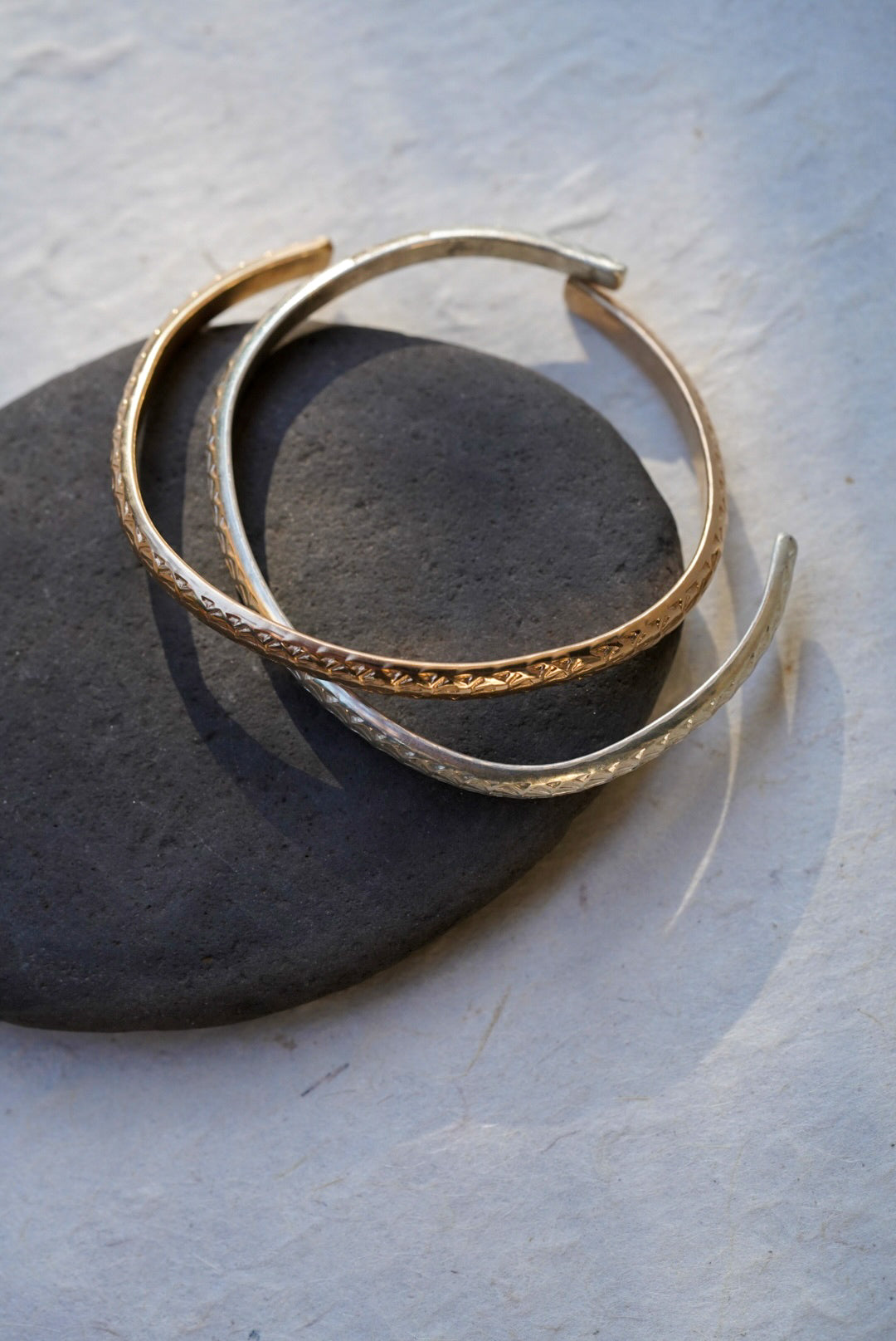 Two textured open cuff bracelets, one gold and one silver, displayed on a dark stone against a light gray background.