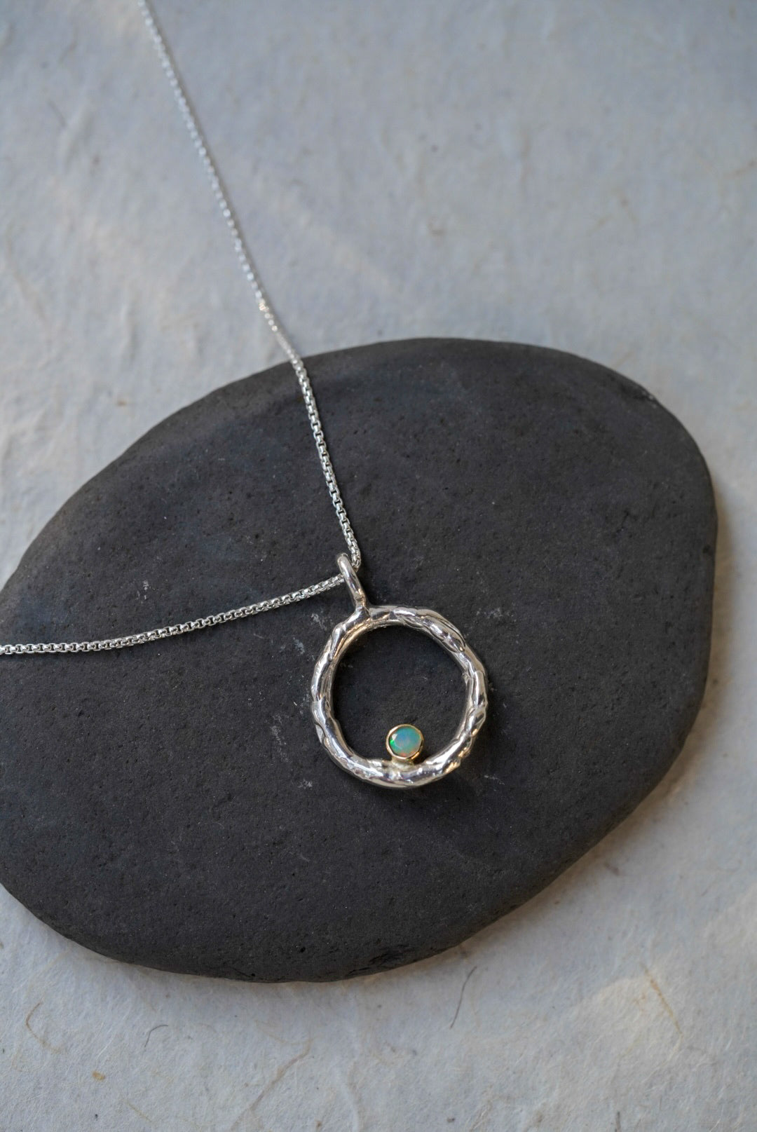 A silver necklace with a textured open circle pendant in a size large and a small opal set in gold, displayed on a dark stone against a light stone background.