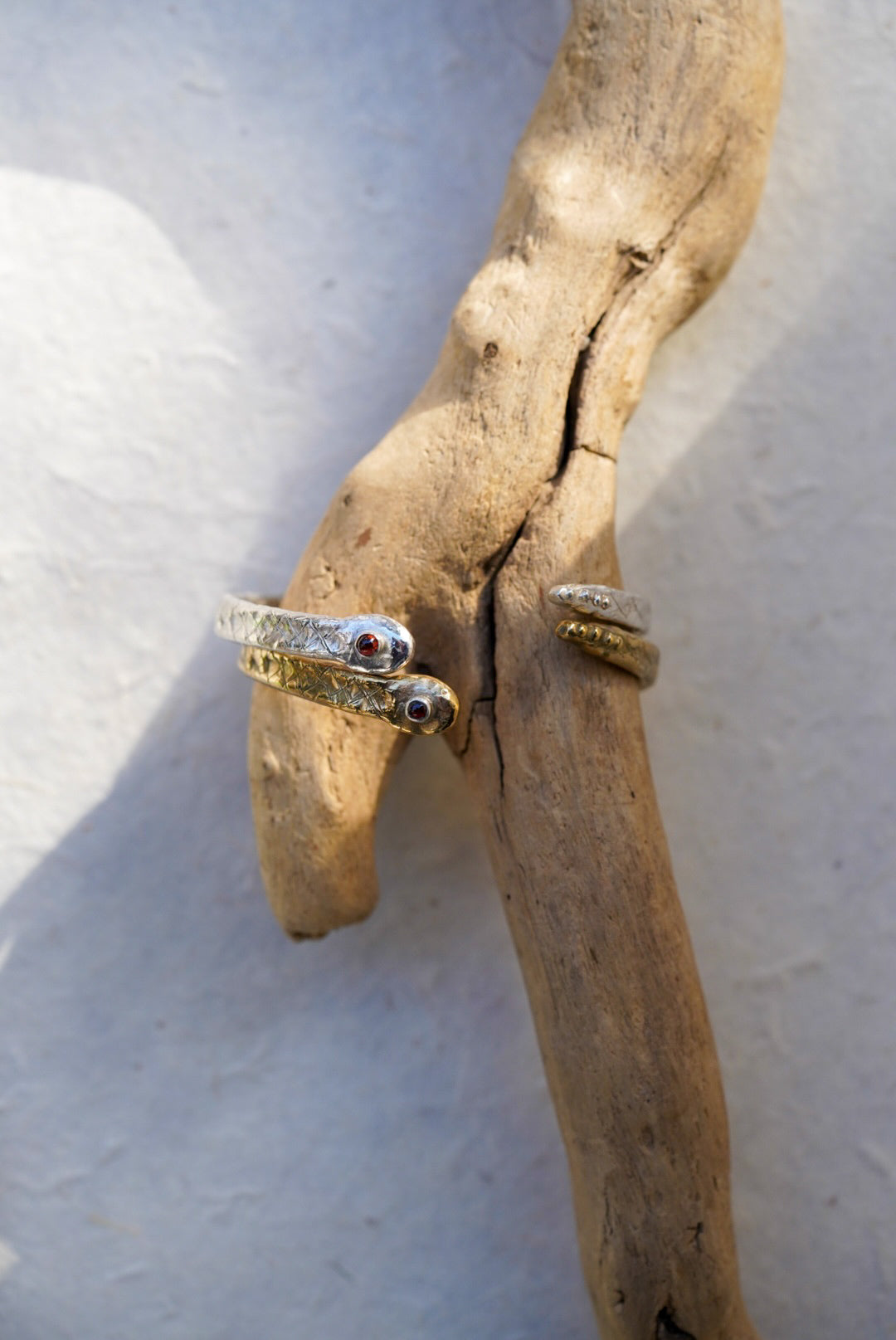 Two snake cuffs around a pice of driftwood, one i9n bronze and one in silver. Both with a garnet in the center of the snakes head. Set on a light background.