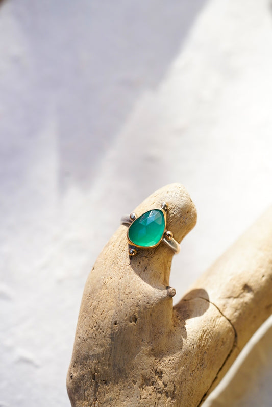 A gold ring with a green gemstone, displayed on a textured beige driftwood piece against a bright, sunlit white background.