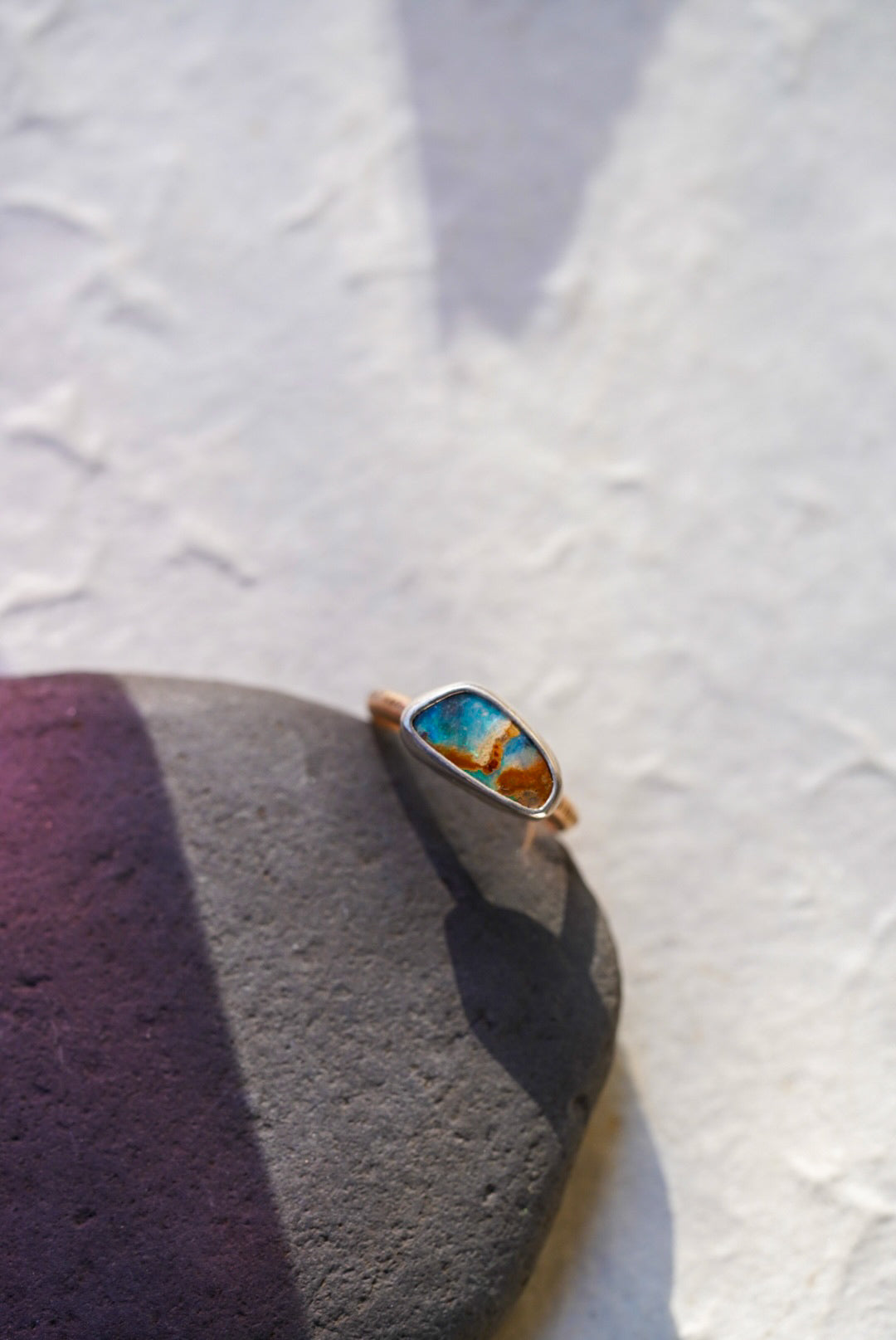 A gold ring with a colorful, marbled turquoise and orange opal, resting on a dark gray stone against a textured white background.