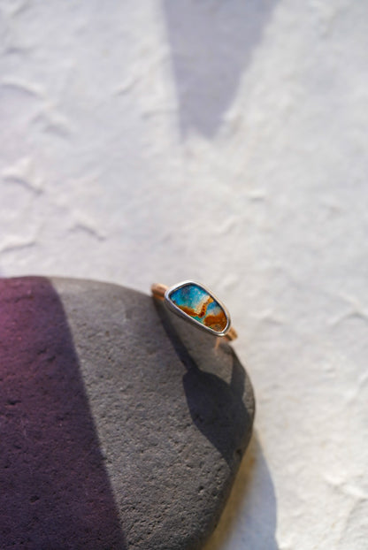 A gold ring with a colorful, marbled turquoise and orange opal, resting on a dark gray stone against a textured white background.