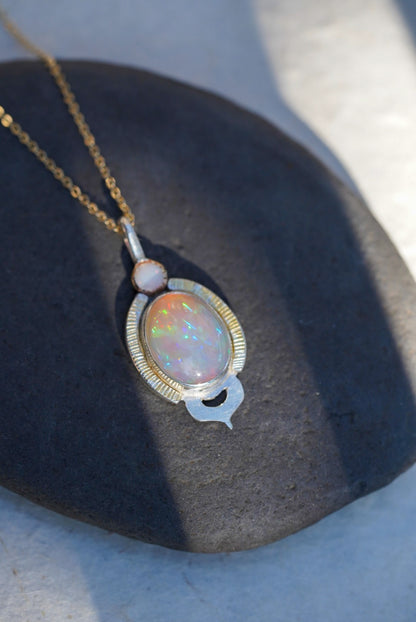 A gold chain necklace with an oval iridescent opal pendant and a small white opal, displayed on a dark stone surface.