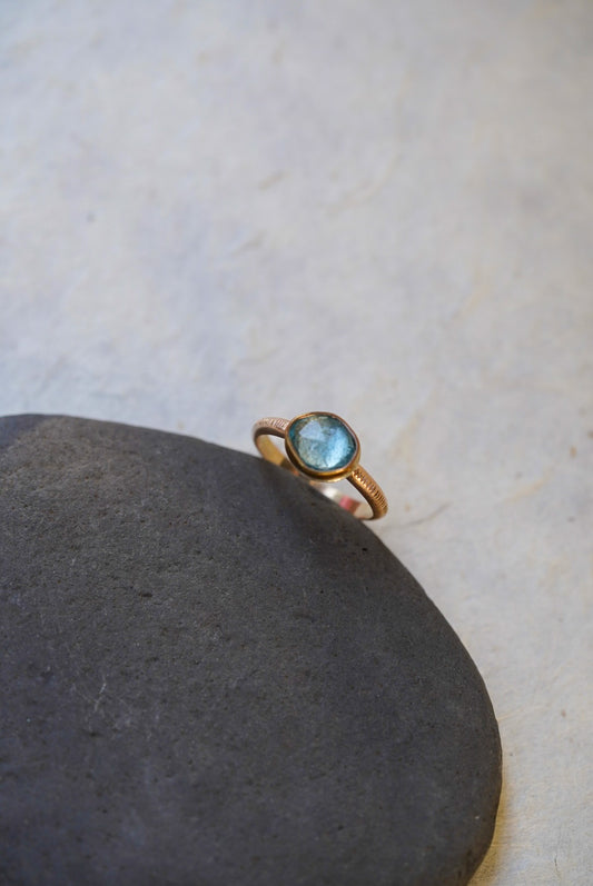 A delicate gold ring with a round light blue gemstone, resting on a smooth dark gray stone against a light beige textured background.