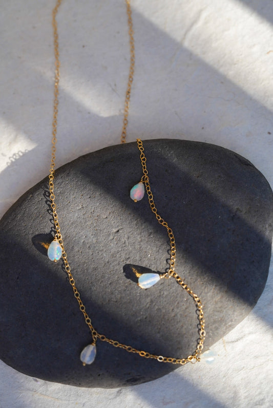 A delicate gold chain necklace with five small, teardrop-shaped iridescent opal beads, draped over a smooth dark gray stone on a textured white surface.