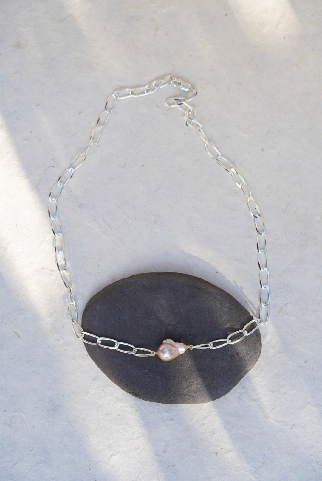 A silver chain necklace with a single irregular light pink pearl pendant, displayed draped over a dark gray stone on a light textured surface.