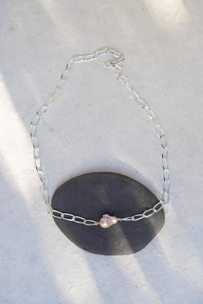A silver chain necklace with a single irregular light pink pearl pendant, displayed draped over a dark gray stone on a light textured surface.