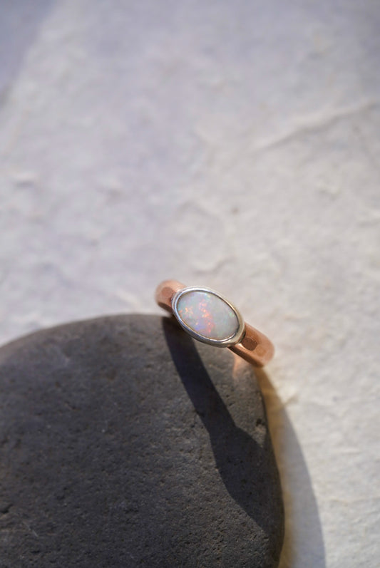 A rose gold ring with an oval-cut opal stone, resting on a dark textured stone against a light, textured background.