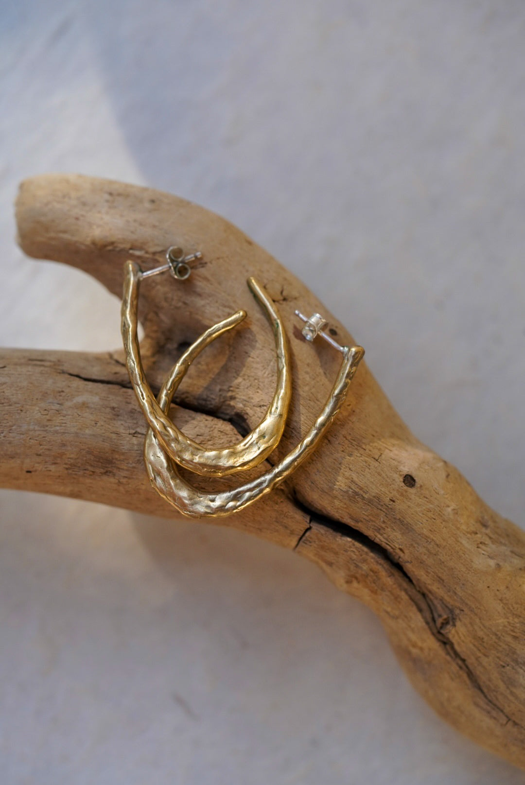 A pair of bronze textured hoop earrings displayed on a piece of natural driftwood against a light gray background.