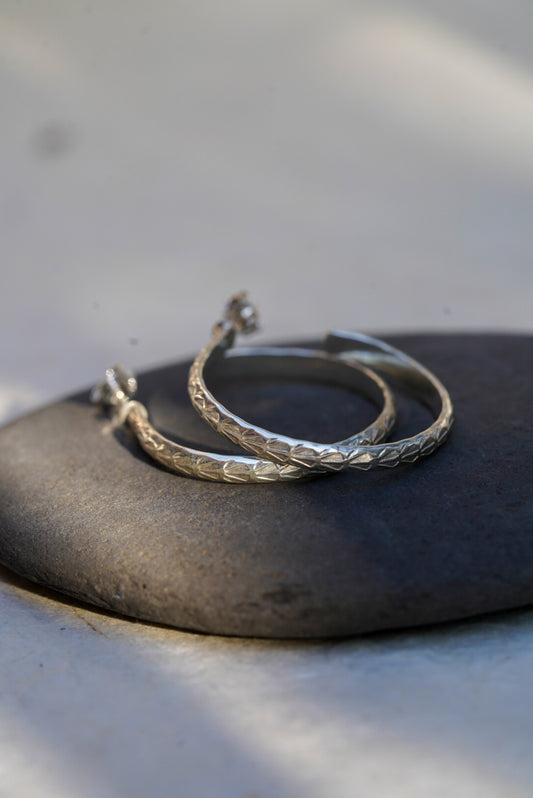A pair of textured sterling silver hoop earrings resting on a smooth dark gray stone, set against a light gray background with soft shadows.