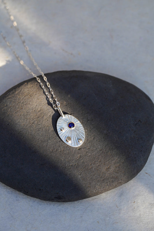A silver oval pendant necklace with a textured sunburst pattern and a small dark blue sapphire, resting on a dark gray stone against a light beige background.