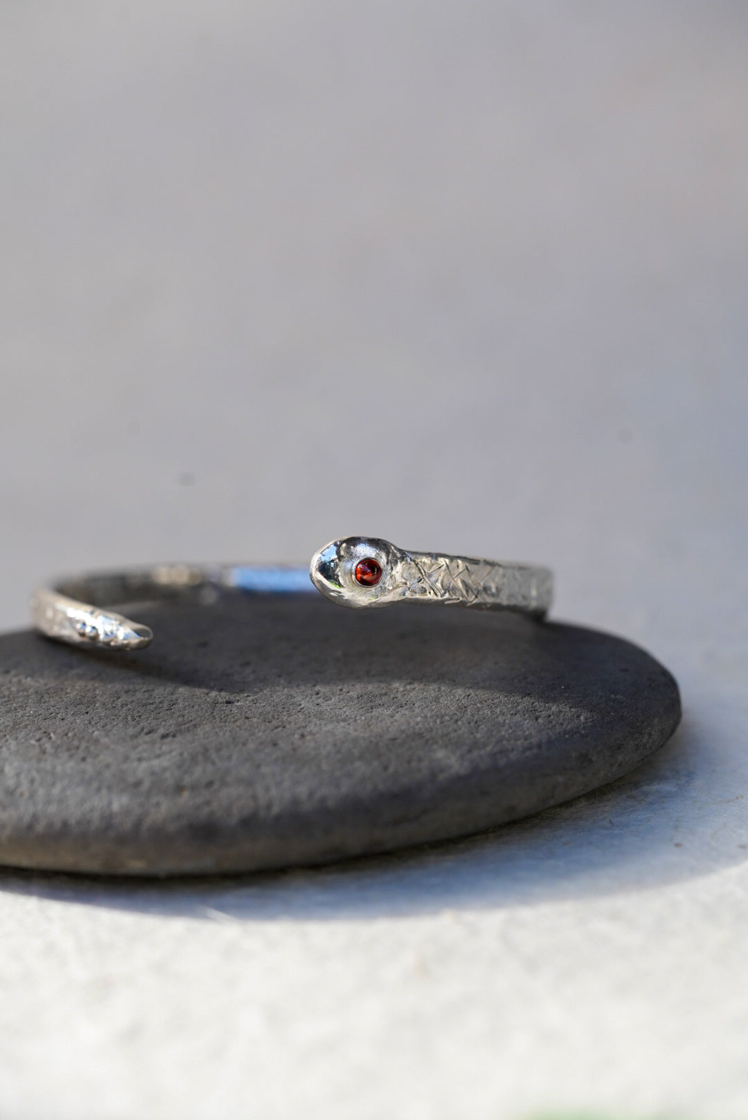 A silver snake cuff with a small garnet, resting on a dark stone against a light gray background.