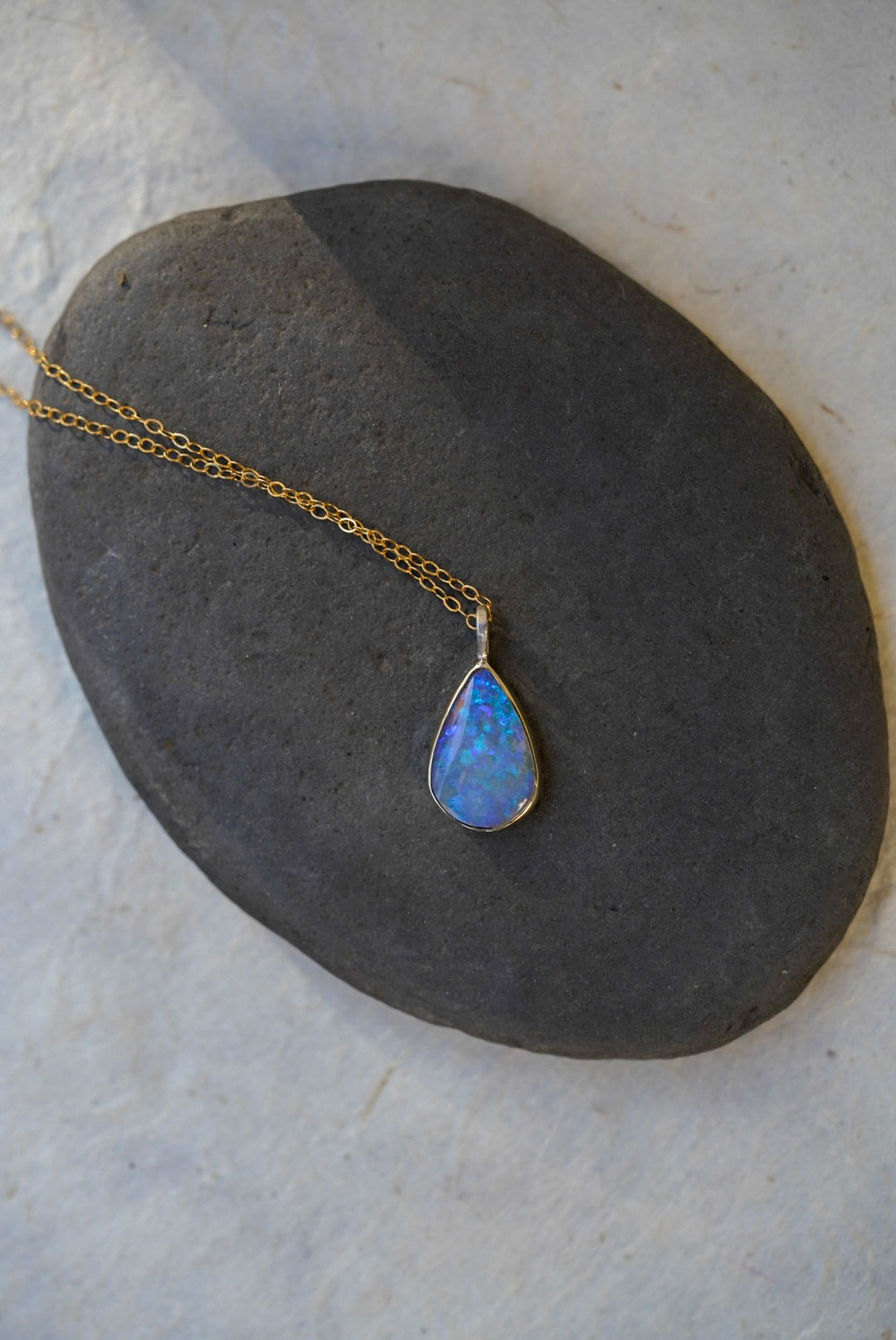A gold chain necklace with a teardrop-shaped blue opal pendant, displayed on a dark gray stone against a light textured background.