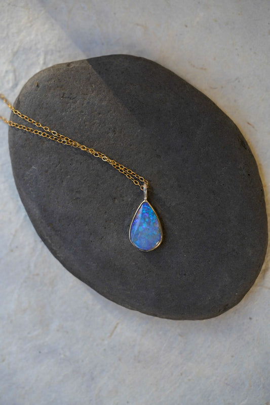 A gold chain necklace with a teardrop-shaped blue opal pendant, displayed on a dark gray stone against a light textured background.