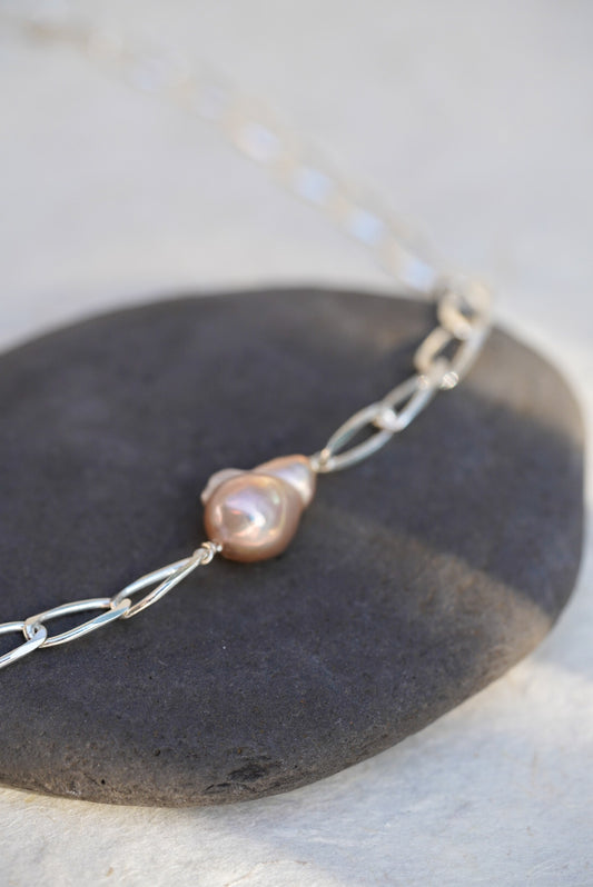 A delicate silver chain necklace featuring a single irregular, lustrous pearl pendant, displayed on a dark gray stone against a light beige background.