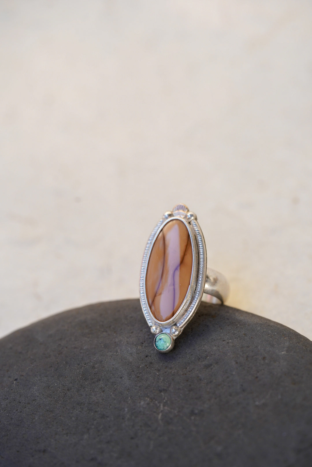 An oval silver ring featuring a brown  stone, accented with a small light green stone, displayed on a dark stone surface against a light beige background.