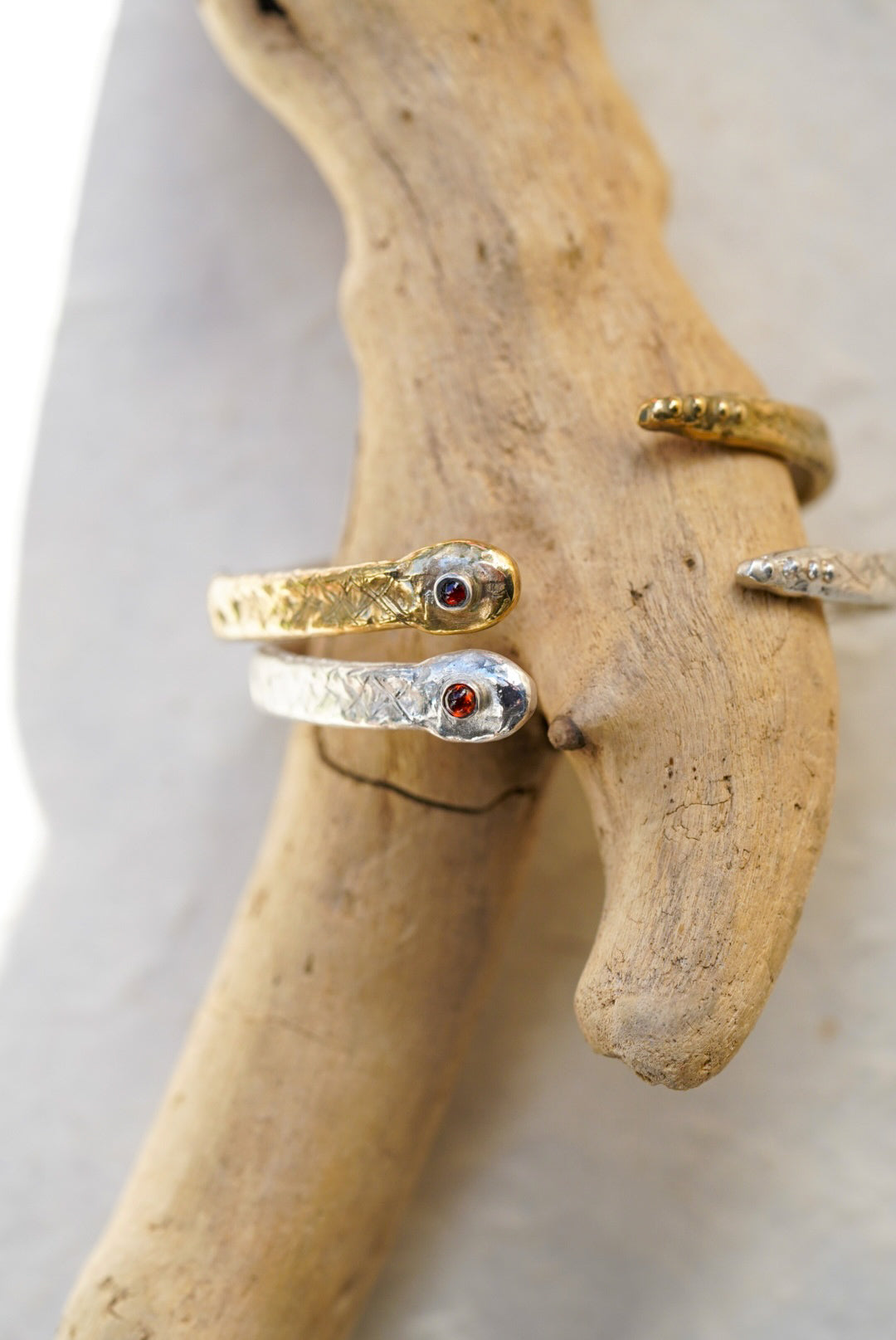 Two snake cuffs around a pice of driftwood, one i9n bronze and one in silver. Both with a garnet in the center of the snakes head. Set on a light background.