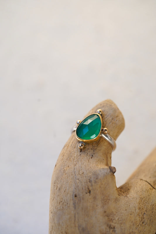 A gold ring with a green gemstone, displayed on a piece of driftwood against a light beige background.