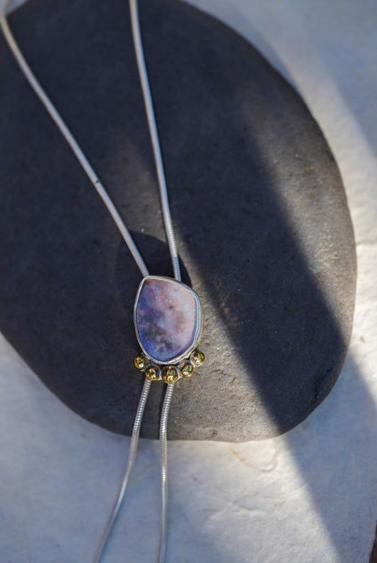A close-up of a silver bolo necklace with a lavender and purple opal stone pendant, accented with small gold accents, resting on a dark gray stone against a light background.