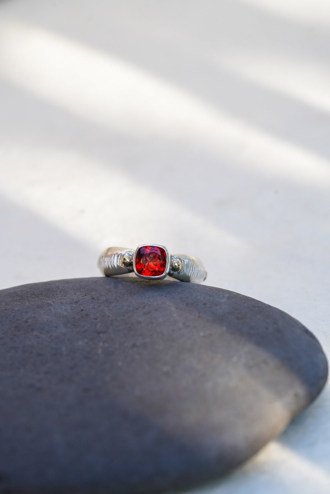 A silver ring with a cushion-cut red gemstone, resting on a smooth dark gray stone against a light background with soft diagonal shadows.