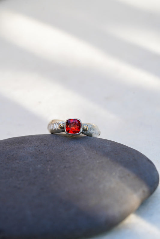 A silver ring with a cushion-cut red gemstone, resting on a smooth dark gray stone against a light background with soft diagonal shadows.