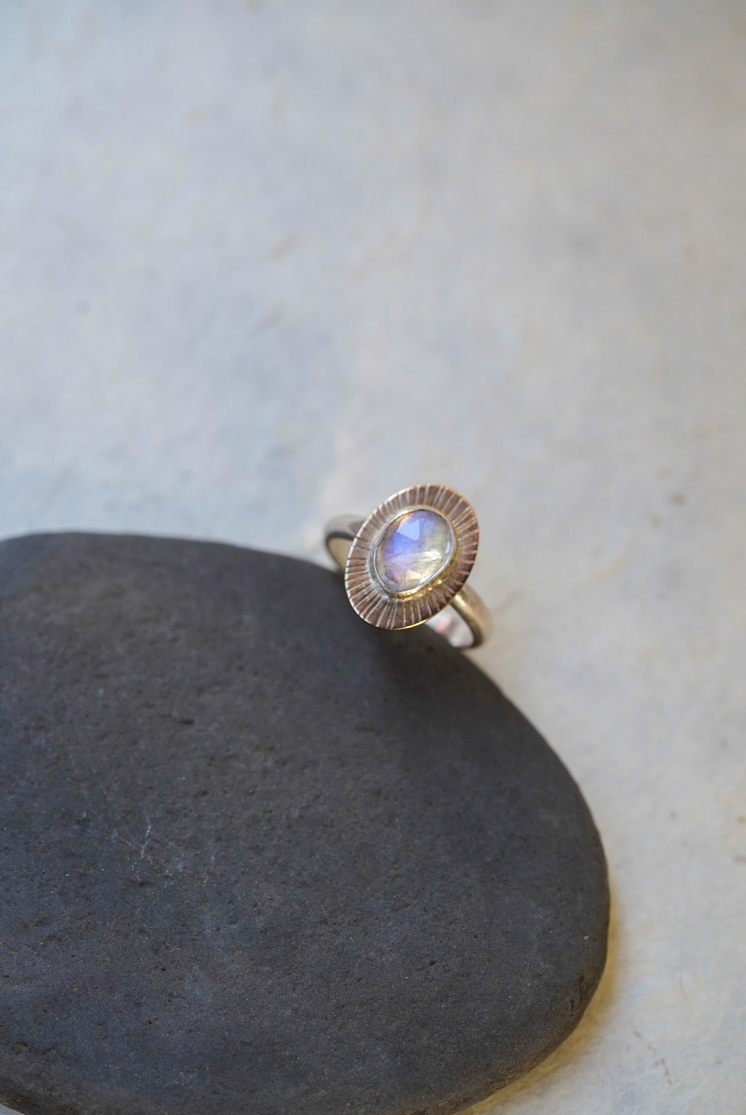 A silver ring with a textured, scalloped oval setting holding a round iridescent moonstone, resting on a dark gray stone against a light gray background.