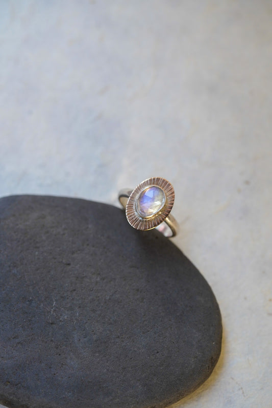 A silver ring with a textured, scalloped oval setting holding a round iridescent moonstone, resting on a dark gray stone against a light gray background.