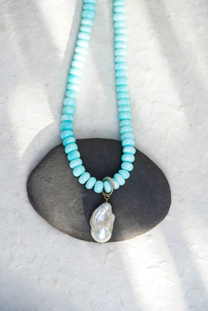 A baby blue beaded necklace with removable charm a large baroque pearl, set upon a black rock on a light background.