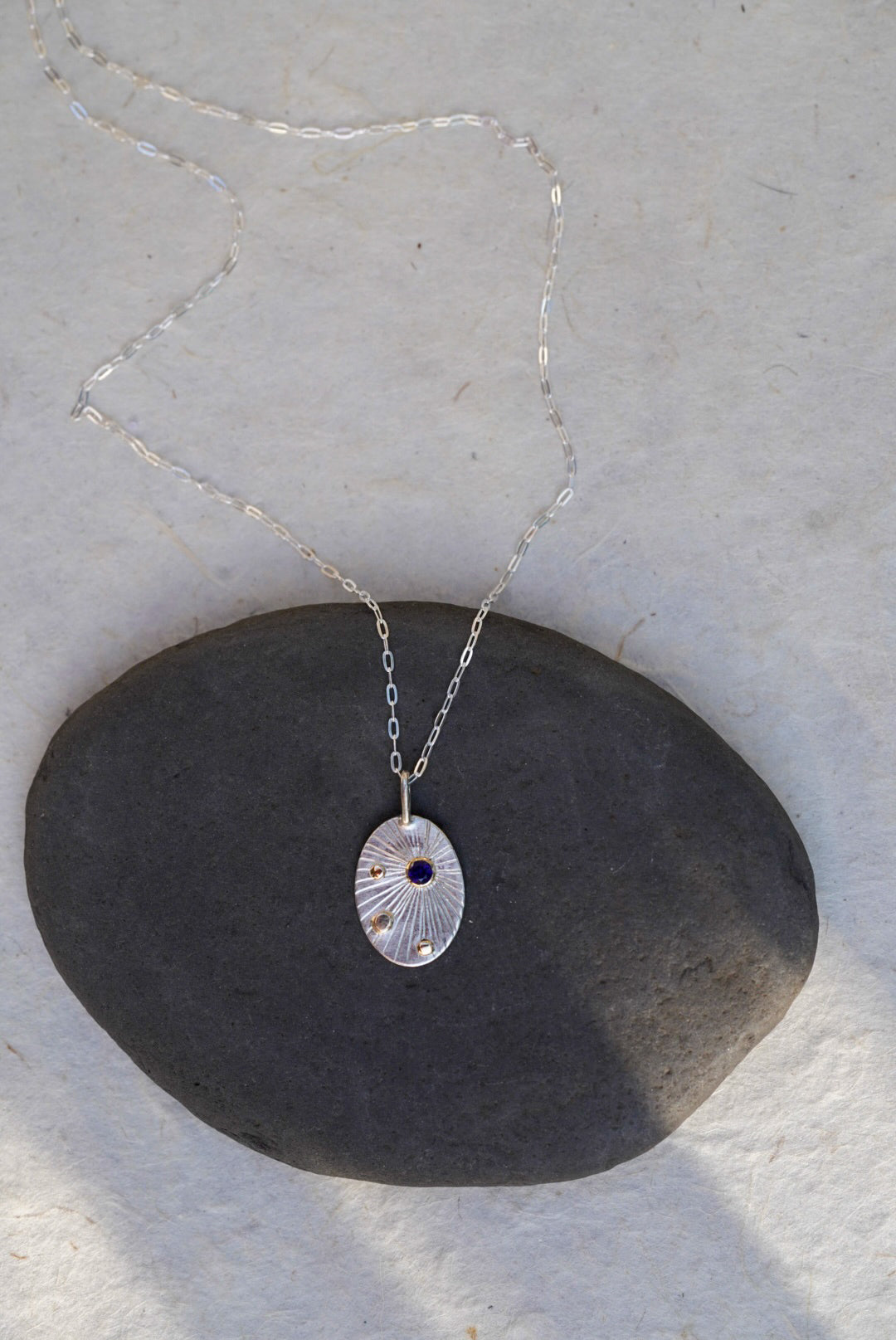 A silver oval pendant necklace with a textured sunburst pattern and a small dark blue sapphire, resting on a dark gray stone against a light beige background.