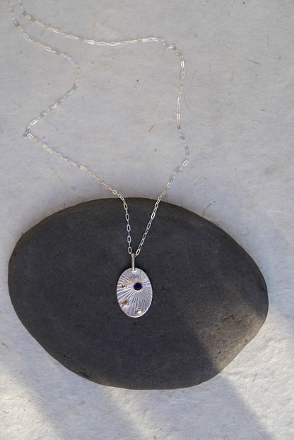 A silver oval pendant necklace with a textured sunburst pattern and a small dark blue sapphire, resting on a dark gray stone against a light beige background.