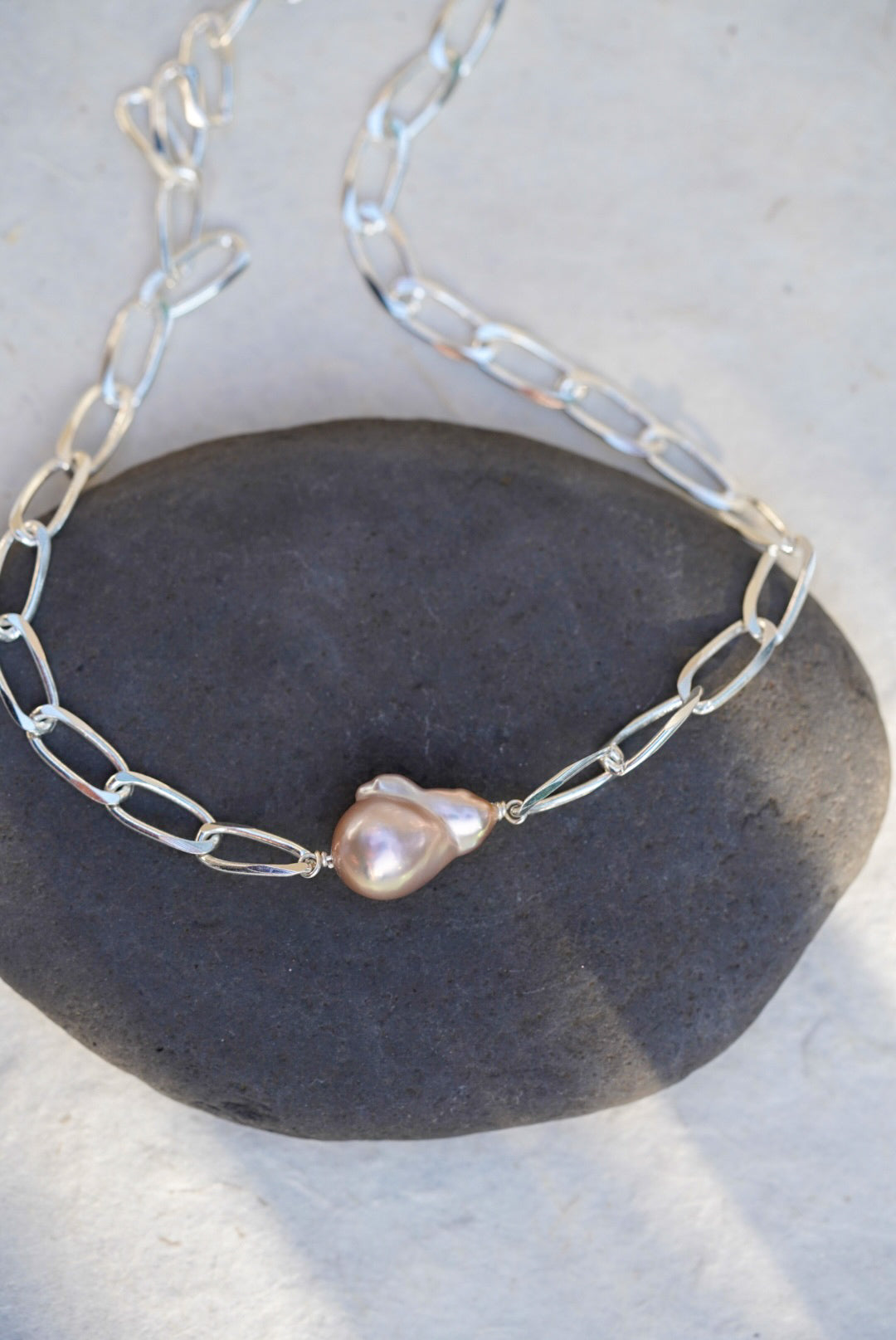 A silver chain necklace with a single irregular baroque pearl pendant, displayed on a dark gray stone against a light gray background.