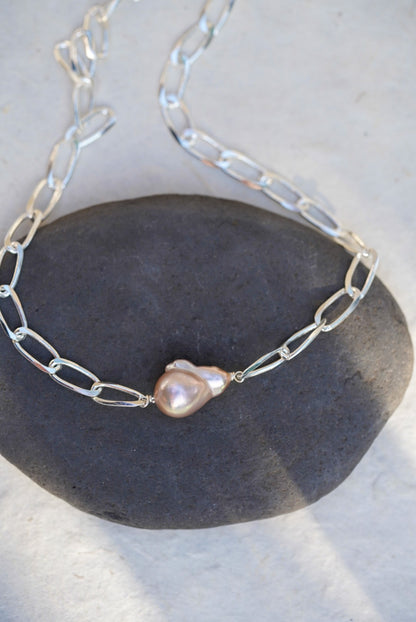 A silver chain necklace with a single irregular baroque pearl pendant, displayed on a dark gray stone against a light gray background.