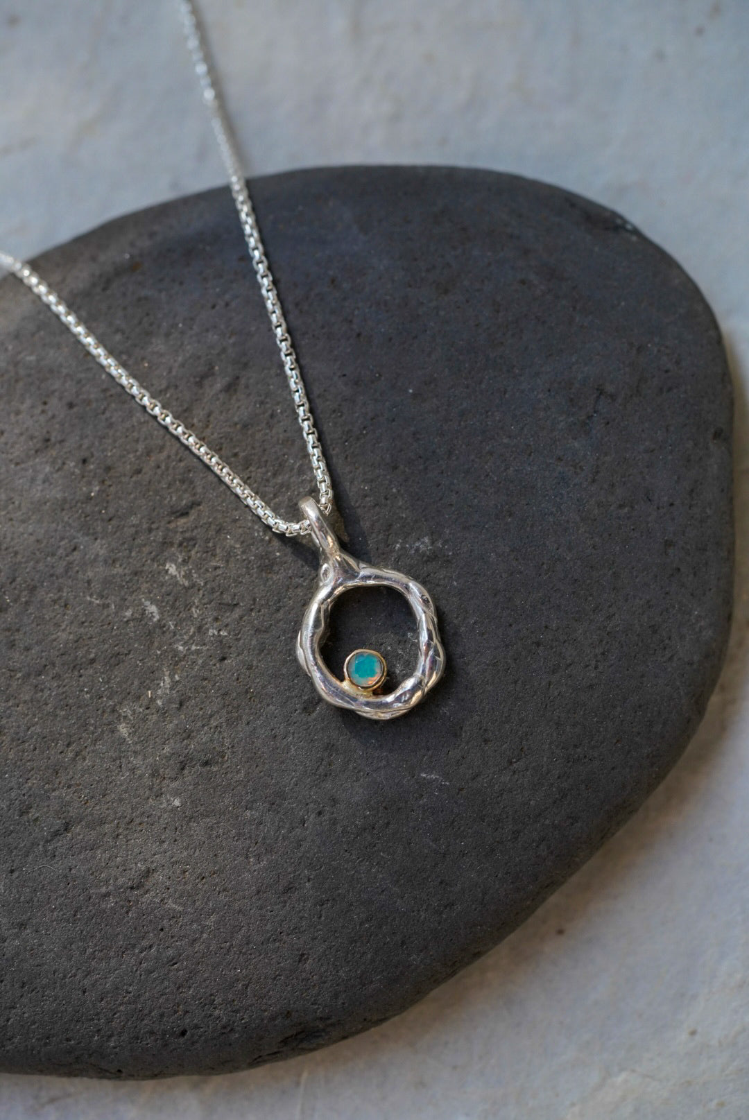 A silver necklace with a textured, organic-shaped ring pendant in size small featuring a small round opal stone, displayed on a dark gray stone surface.