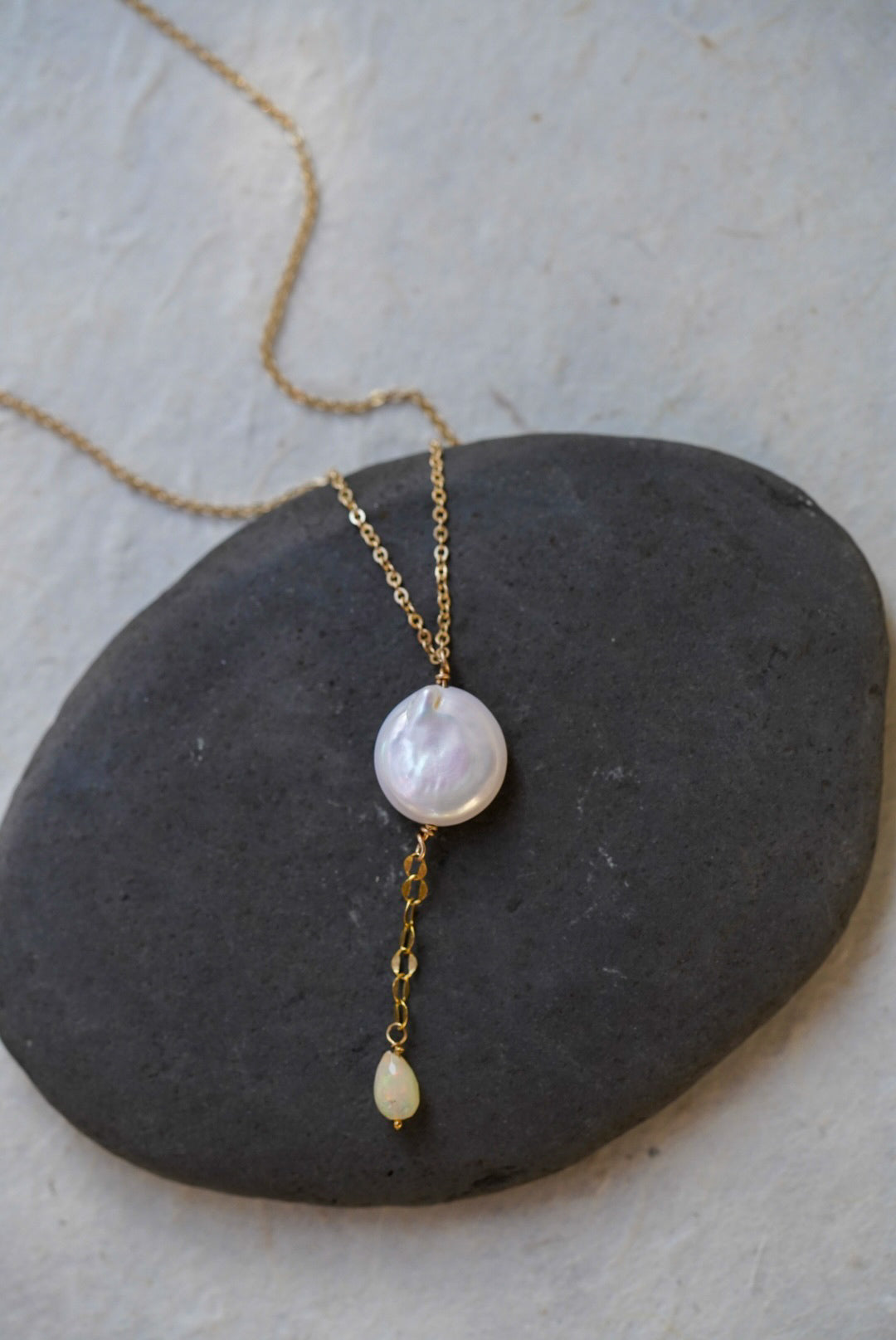 A gold chain necklace with a round iridescent white pearl pendant and a small gold teardrop opal, displayed on a dark stone over a light textured background.